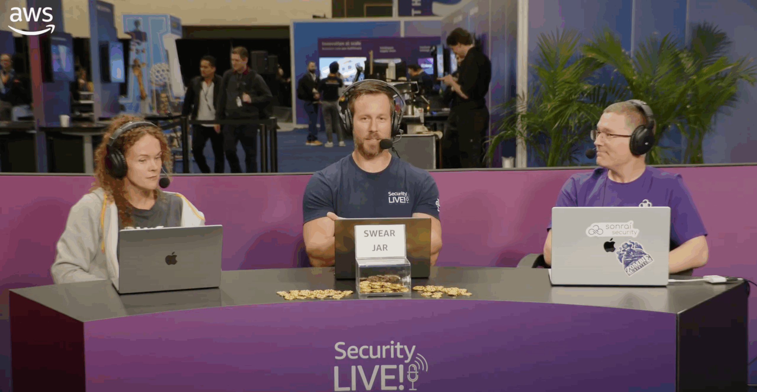 Three people wearing headsets sit behind a table with laptops and a box labeled "Swear Jar" filled with coins, at the "Security LIVE!" event. Purple banners and a busy exhibition area are in the background.